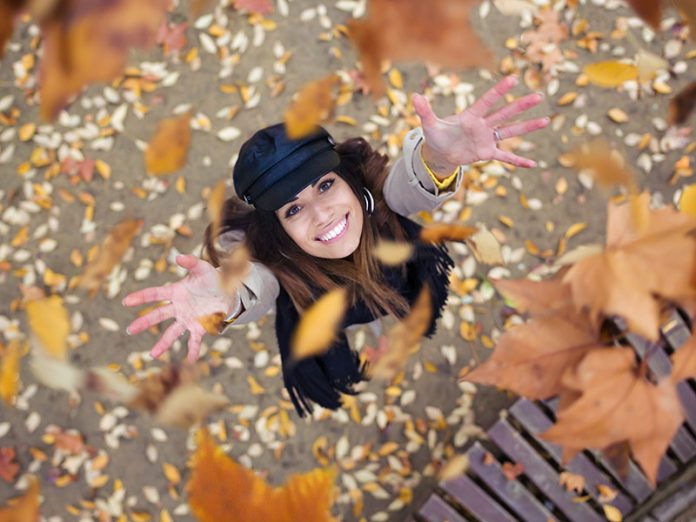 Pretty young woman looking to the sky with arms raised as leaves fall from the trees in the park in autumn.