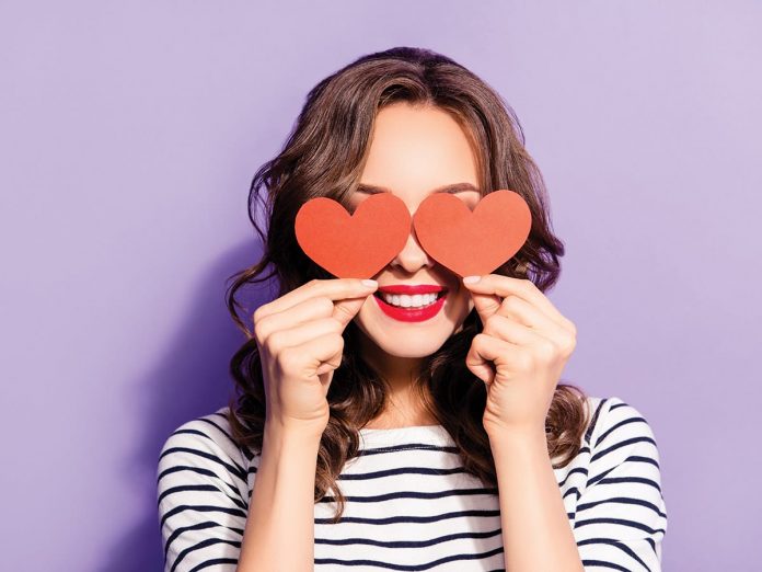 Portrait of lovely creative girl with white teeth red pomade covering closing eyes with two carton paper small little heart figures love signs isolated on violet background