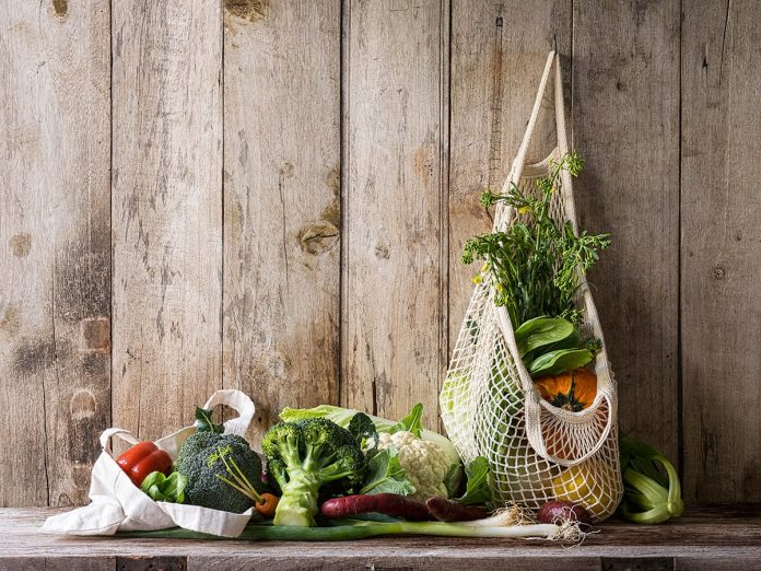 Market fresh vegetables hanging in a reusable string cotton bag, on an old wood board wall background, above an old wooden table with other vegetables.
